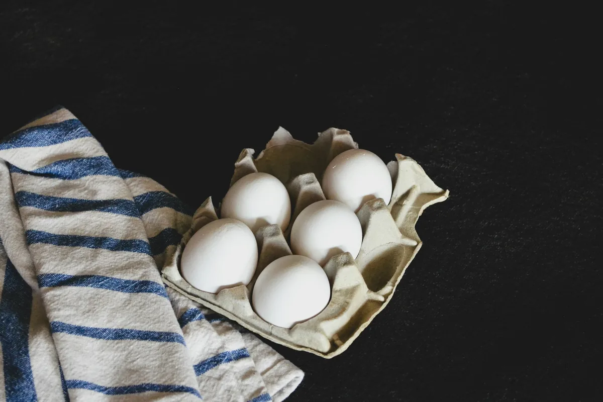 Five white eggs in a cardboard egg tray beside a striped kitchen towel on a dark table