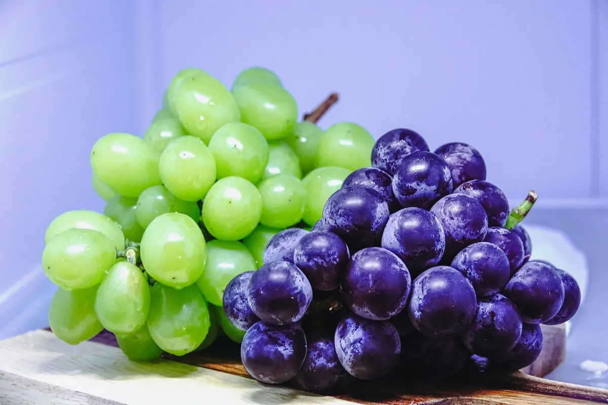 A bunch of green grapes and a bunch of dark purple grapes on a wooden cutting board