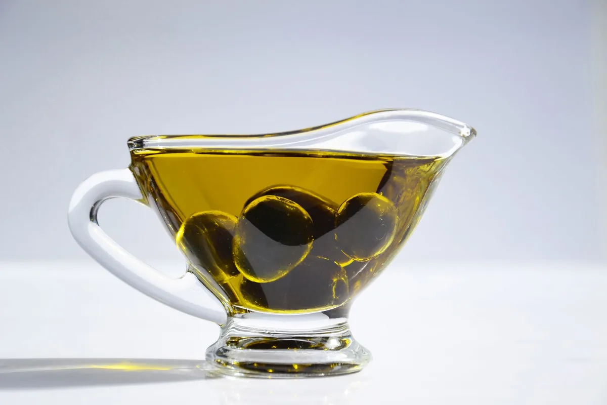 A glass gravy boat of golden olive oil with dark olives inside, on a white surface