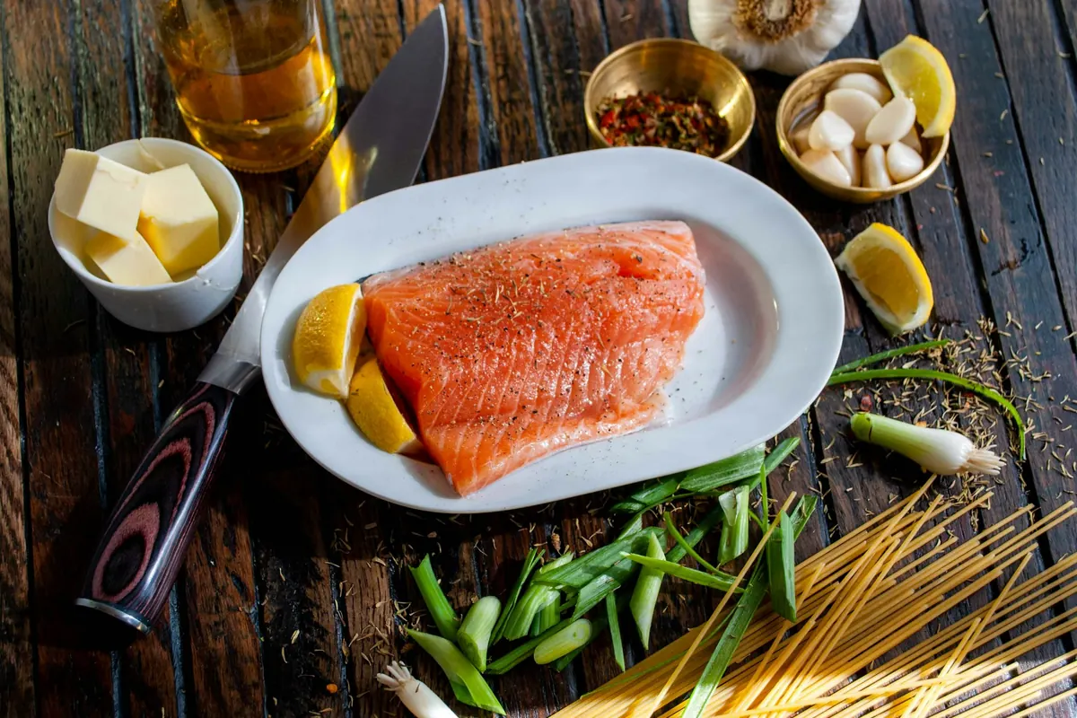 A raw salmon fillet with lemon wedges on a white plate, surrounded by garlic, spring onions and spices on a wooden table