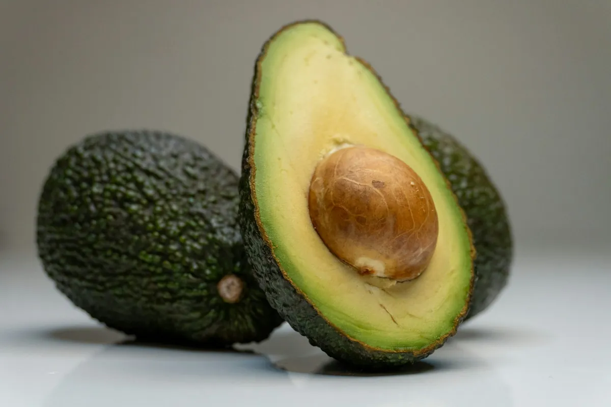 A halved avocado with its stone leaning against a whole avocado