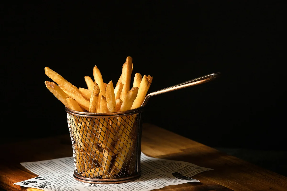 Golden French fries in a small wire fry basket on a wooden board against a dark background